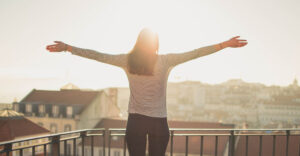 Eine Frau steht auf dem Balkon. Die Morgensonne scheint. Sie hällt die Arme hoch und freut sich.